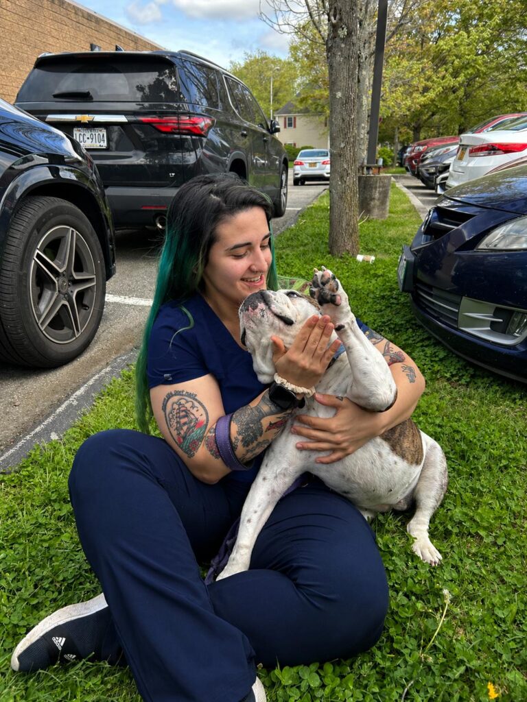 Veterinary technician with colorful hair holding happy brindle and white dog outdoors at animal hospital